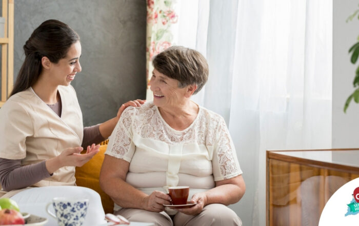 Smiling caregiver with her hand on an older woman’s shoulder as they share tea on the couch, showing home care vs assisted living