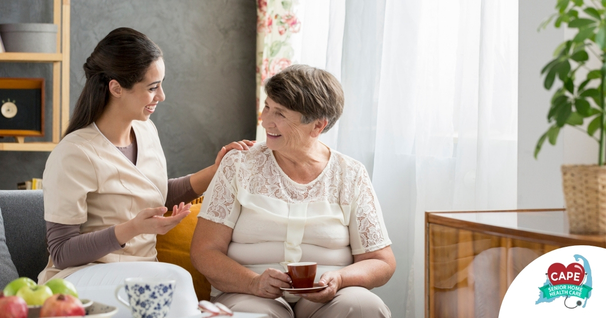 Smiling caregiver with her hand on an older woman’s shoulder as they share tea on the couch, showing home care vs assisted living